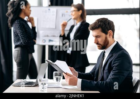 Busy focused business man in a formal suit, sit at workplace in the office, looks out the documents, financial reports. On the background his female colleagues discussing financial strategy Stock Photo