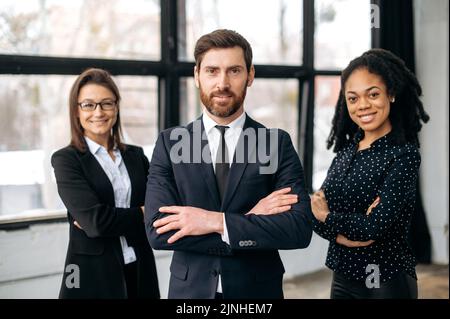 Successful business partners. Multiracial successful colleagues, dressed in formal wear, standing in their modern office with their arms crossed, looking at the camera, smiling friendly Stock Photo