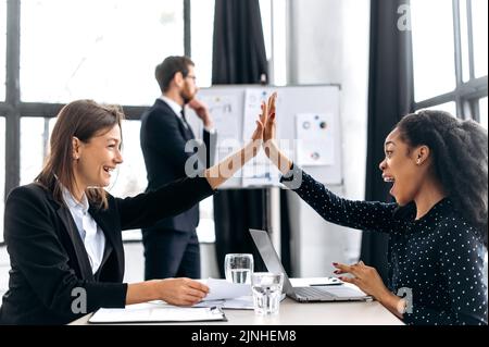 Two successful multi-ethnic female colleagues work in a modern office on a project, give each other five, rejoice at a successful deal, good profit, a successful startup, smiling. Agreement, deal Stock Photo