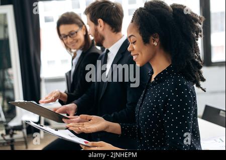 Side view to successful multiracial people, business partners, are standing in their modern office, happily discussing the project, predicting its success, discussing new ideas, smiling Stock Photo