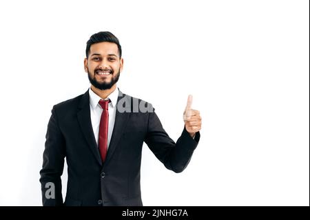 Successful handsome confident indian or arabian businessman in a black business suit, standing over isolated white background, showing thumb up, agree sign, looking at camera, smiling happily Stock Photo
