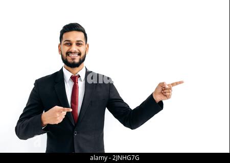 Friendly positive confident indian or arabian businessman in a black business suit, standing over isolated white background, pointing fingers to the side, looking at camera, smiling happily Stock Photo