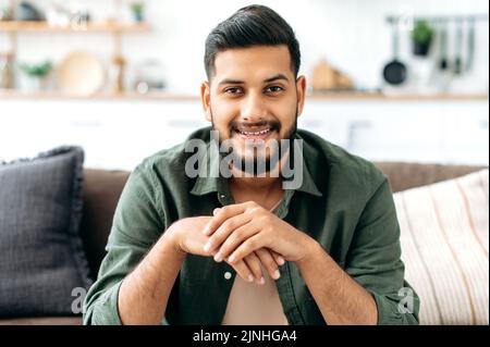 Portrait of confident attractive indian or arabian man, in casual stylish clothes, sitting at home in cozy living room on the background of the kitchen, looks at camera, smiling friendly Stock Photo