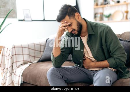 Unhappy indian or arabian man, sits on comfortable sofa in cozy living room, holds his hands on his stomach, grimaces from pain in his stomach, suffers from poisoning, spasm, stomach problems Stock Photo
