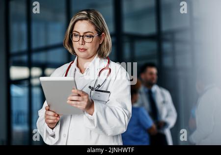 Connected and on call. a young doctor using a digital tablet in a hospital with her colleagues in the background. Stock Photo