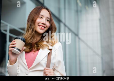 Asian businesswoman holding coffee cup takeaway going to work she walking near her office building, Portrait smiling business woman hold paper cup of Stock Photo