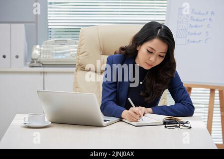 Concentrated Asian businesswoman with long curly hair sitting at office desk and taking notes while preparing for important negotiations, portrait shot Stock Photo