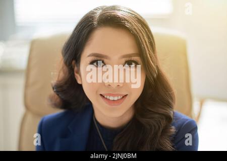 Head and shoulders portrait of beautiful Asian businesswoman looking at camera with charming smile while sitting at office desk Stock Photo