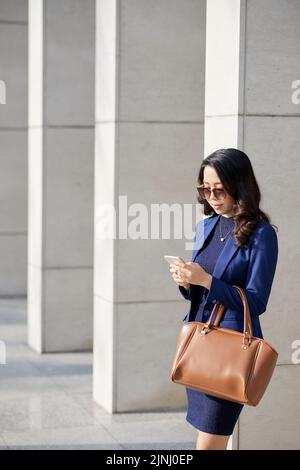 Pretty Asian designer wearing sunglasses standing at facade of office building and texting with friend on smartphone, portrait shot Stock Photo