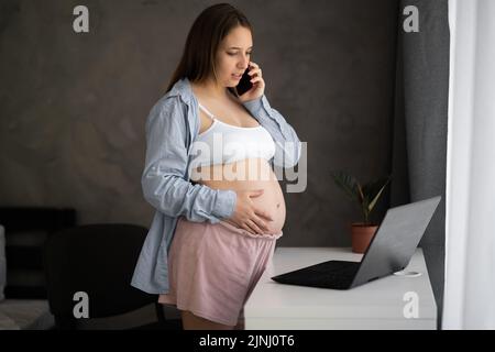 Beautiful pregnant woman is talking on mobile phone, holding one hand on her belly and smiling while standing near working place at home, using laptop Stock Photo