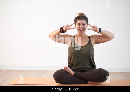 Attractive young woman wearing leggings and top showing victory gesture while sitting in lotus position at modern health club, copy space Stock Photo