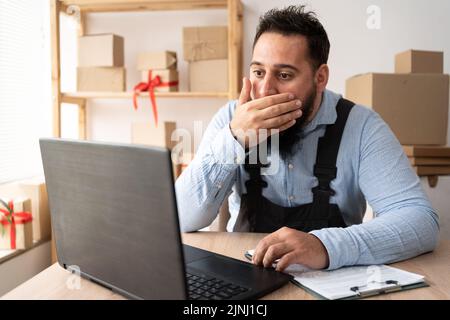 Nervous Indian man close mouth read amazing news on laptop computer while working remotely, stressed guy shocked with sales in web store for devices Stock Photo