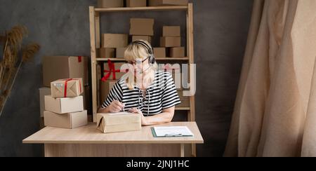 Happy older businesswoman in headset talking on conference call while placing order in warehouse, mature aged woman, call center agent, start up Stock Photo