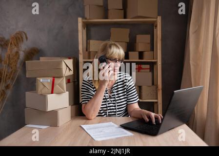 Older female dropshipping business owner using laptop talking on phone in warehouse. Mature woman seller merchant checking online order, e-commerce Stock Photo