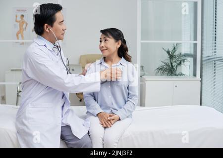 Profile view of confident middle-aged therapist wearing white coat using stethoscope while examining senior patient, interior of modern ward on background Stock Photo