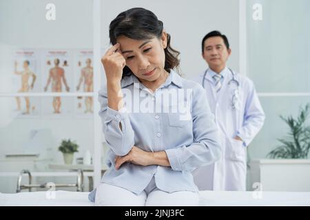 Sad Asian patient sitting on bed of modern ward and looking down while highly professional doctor wearing white coat standing behind her Stock Photo
