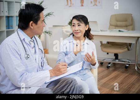 Pretty aged woman sitting on cozy sofa and describing medical symptoms to confident physician while having consultation at modern office Stock Photo