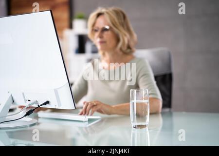Water Drinking Glass On Desk And Woman In Foreground Using Computer Stock Photo