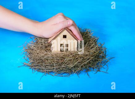 Hand covers a miniature wooden house placed in a hay nest isolated on blue background. Home insurance concept. Stock Photo