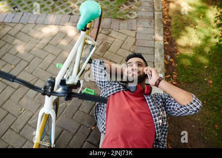 High angle view of cheerful Indian man talking to friend on smartphone while taking rest from cycling, he lying on park bench while bicycle standing next to him Stock Photo
