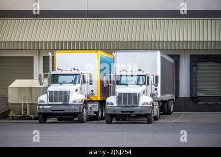 Industrial standard for local deliveries and short haul freights compact middle duty rigs semi trucks with box trailers standing in warehouse dock wit Stock Photo