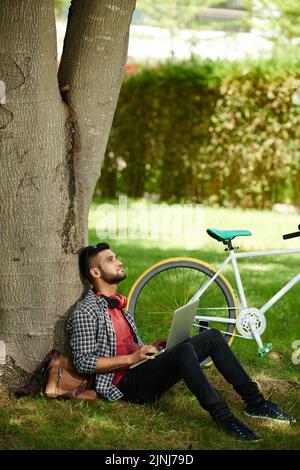 Pensive young freelancer sitting on green lawn of public park and working on promising project with help of laptop, bicycle standing next to him Stock Photo
