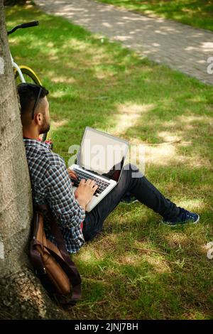 Back view of bearded young freelancer leaning on tree while finishing ambitious project with help of laptop, green public park illuminated with sunbeams on background Stock Photo