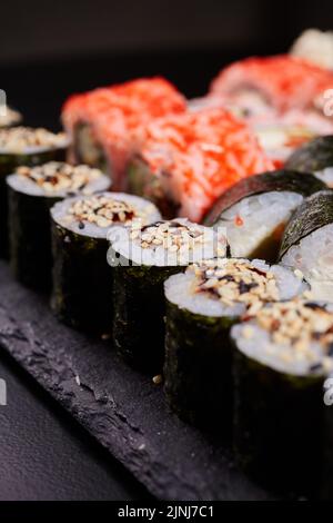 Close up of sushi with sesame seeds wrapped in nori leaves lying on a black board on the background of other types of sushi. Blurred background. Selec Stock Photo