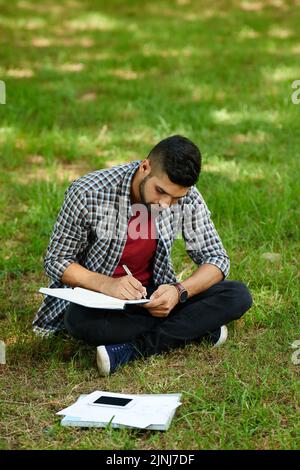 Concentrated Indian student in casualwear sitting in lotus position on green lawn and taking notes while preparing for university exam Stock Photo