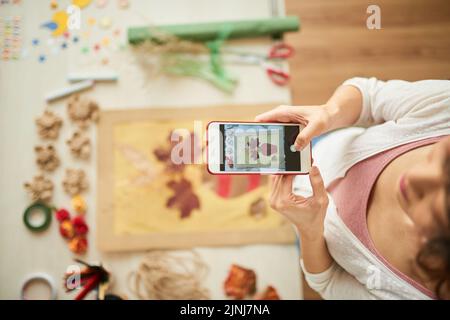 High angle view of young woman in casualwear standing at wooden table and taking picture of just finished handmade decor, focus on foreground Stock Photo