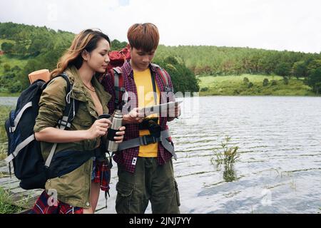 Loving Asian couple studying map on modern digital tablet while taking break from hiking, pretty young girl holding vacuum flask in hands, picturesque view on background Stock Photo