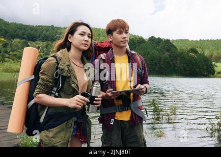 Two Asian hikers with backpacks enjoying picturesque view while standing on shore of lake and warming with hot tea, handsome man using digital tablet in order to check map Stock Photo