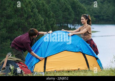 Cheerful Asian tourists having fun while setting up tent in picturesque place, green forest and deep lake on background Stock Photo