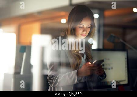 Business woman reading message on phone, checking notifications and browsing online for a cab after working late in dark office. Corporate Stock Photo
