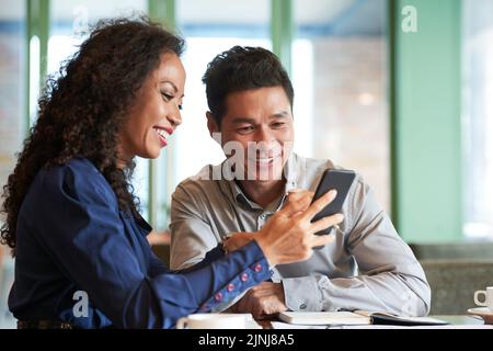 Smiling middle-aged colleagues having fun while sitting at cozy small coffeehouse and analyzing results of accomplished work, pretty woman showing something on smartphone Stock Photo
