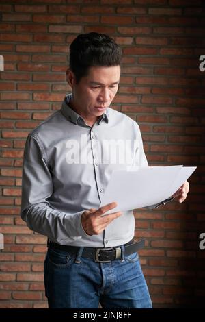 Portrait shot of confident middle-aged businessman wearing jeans and shirt studying documents while standing at brick wall Stock Photo