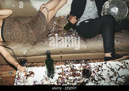 Home party at full speed: unrecognizable young man and women relaxing on sofa with alcoholic drinks in hands, colorful confetti scattered everywhere Stock Photo