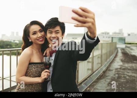 Attractive Vietnamese woman with glass of red wine in hand posing for photography while her smiling boyfriend taking selfie on smartphone, picturesque cityscape on background Stock Photo