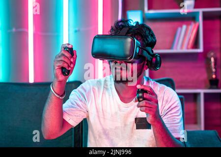 Focus on VR, Young gamer playing video game on virtual reality or VR headset using joystick at home - concept of metaverse, cyberspace and futuristic. Stock Photo