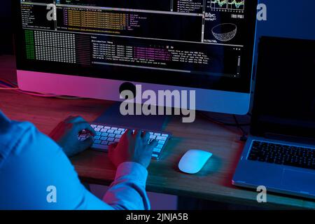 Close-up shot of unrecognizable programmer wearing white shirt sitting in front of modern computer and writing programming code at dim office Stock Photo