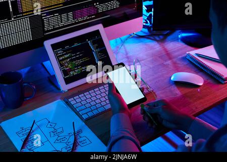 Unrecognizable coder using modern smartphone while sitting at office desk and working on ambitious start-up project, close-up shot Stock Photo