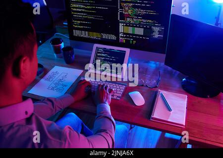 Over shoulder view of hard-working programmer wearing shirt sitting in front of computer and writing code at dim office Stock Photo