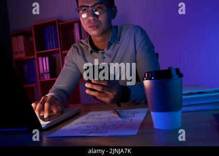Portrait of concentrated Asian manager in eyeglasses sitting in front of laptop and finishing annual accounts while working late in dim office Stock Photo