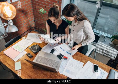Top view of two female colleagues and desk covered with papers and documents. Businesswomen studying the data working on new sales strategy. Stock Photo