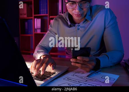 Handsome Asian programmer wearing shirt and eyeglasses putting finishing touches in brand new computer app while sitting at desk in dim office, waist-up portrait Stock Photo