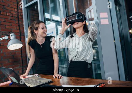 Two female application developers testing a new app designed for VR headset standing in modern office. Stock Photo