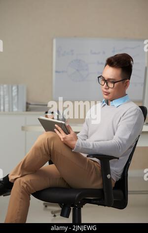 Young confident businessman sitting in armchair and reading data on tablet screen Stock Photo