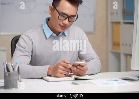 Young Vietnamese businessman smiling when reading text message in his smartphone Stock Photo