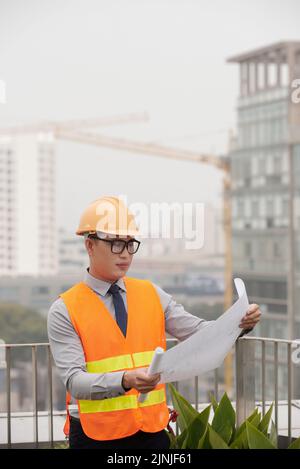 Portrait of young Asian architect analyzing construction plan in his hands Stock Photo