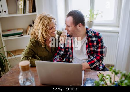 Young man with Down syndrome sitting at desk in office and using laptop, his mother helping him. Stock Photo